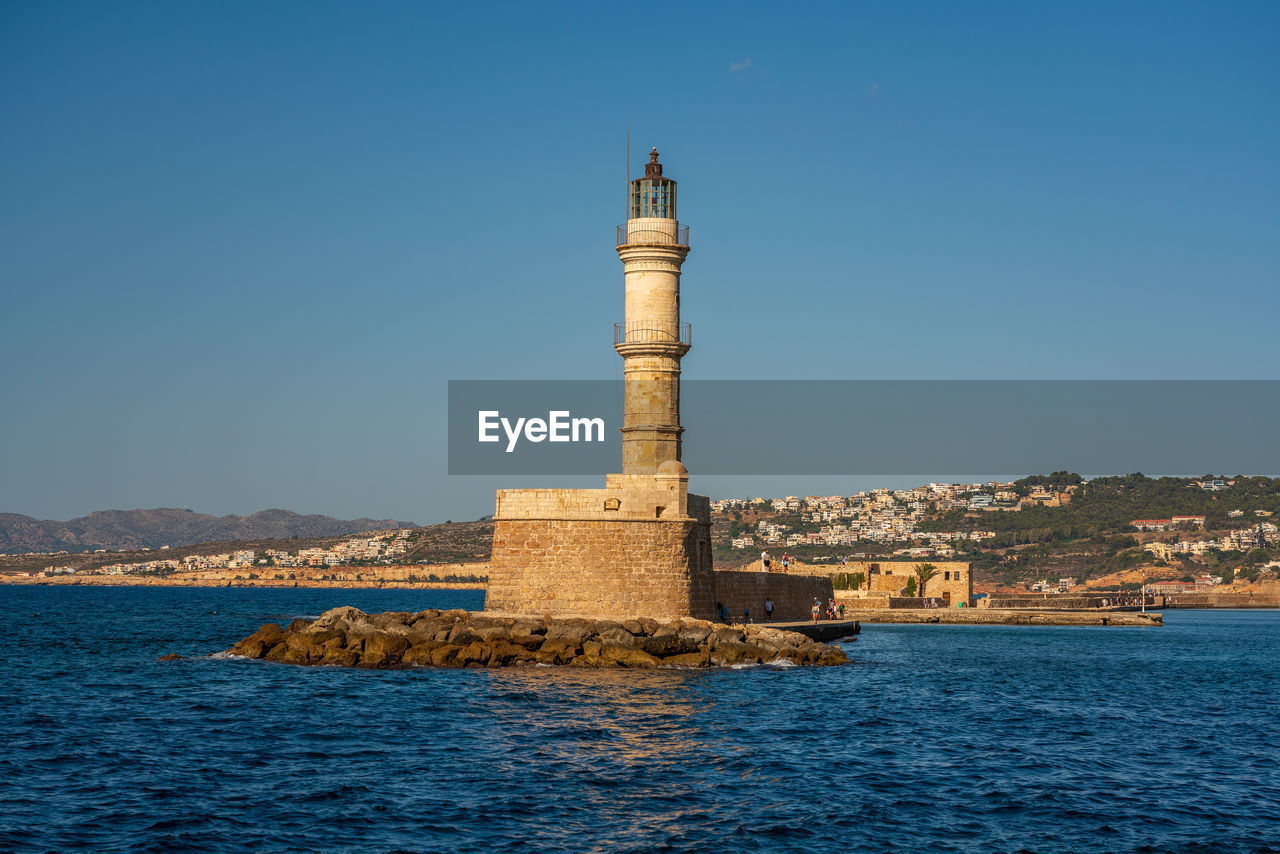 Lighthouse by sea against clear blue sky