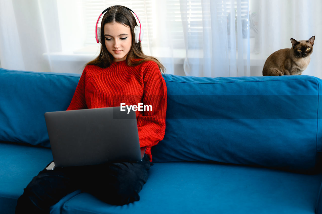 A young girl in a red  sweater works on a laptop at home. her cat and dog are sitting next to her