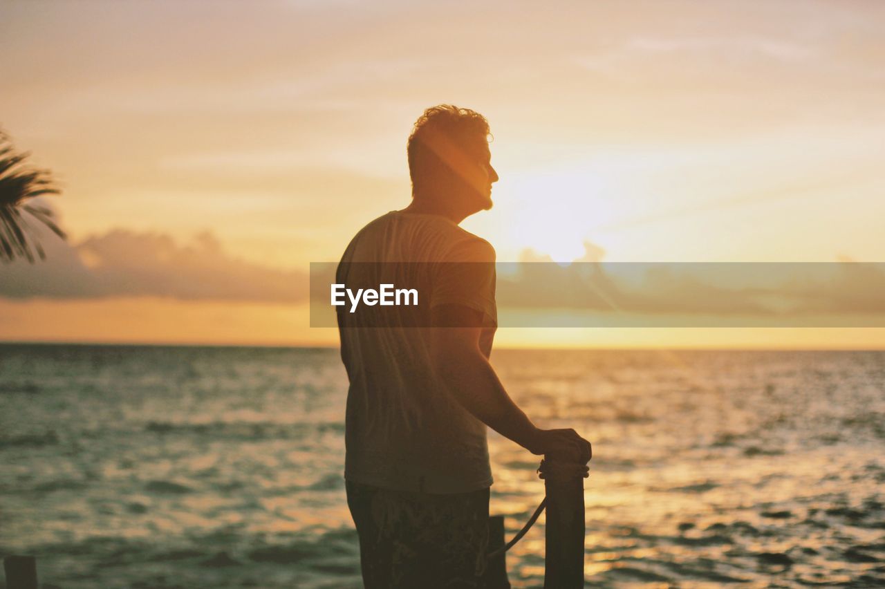 Rear view of young man looking at sea while standing against sky during sunset