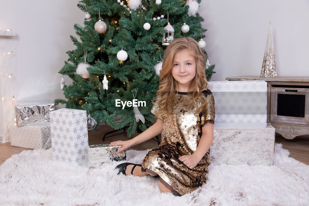 Portrait of girl sitting on carpet with gift by christmas tree at home