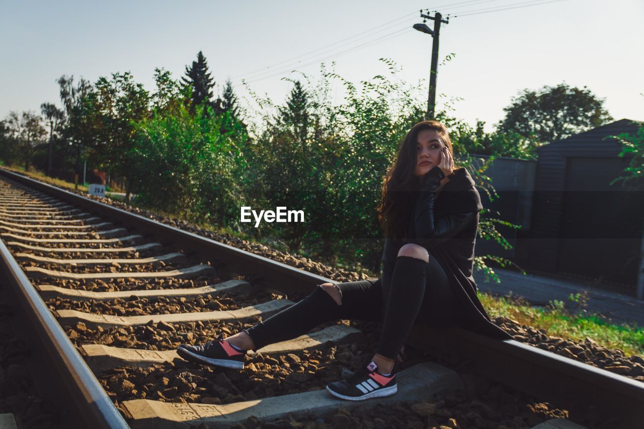 PORTRAIT OF YOUNG WOMAN SITTING ON RAILROAD TRACK AGAINST TREES