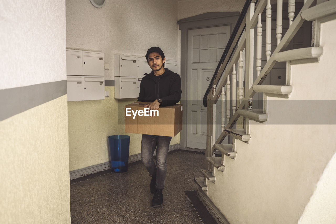 Full length of delivery man with package box walking by staircase