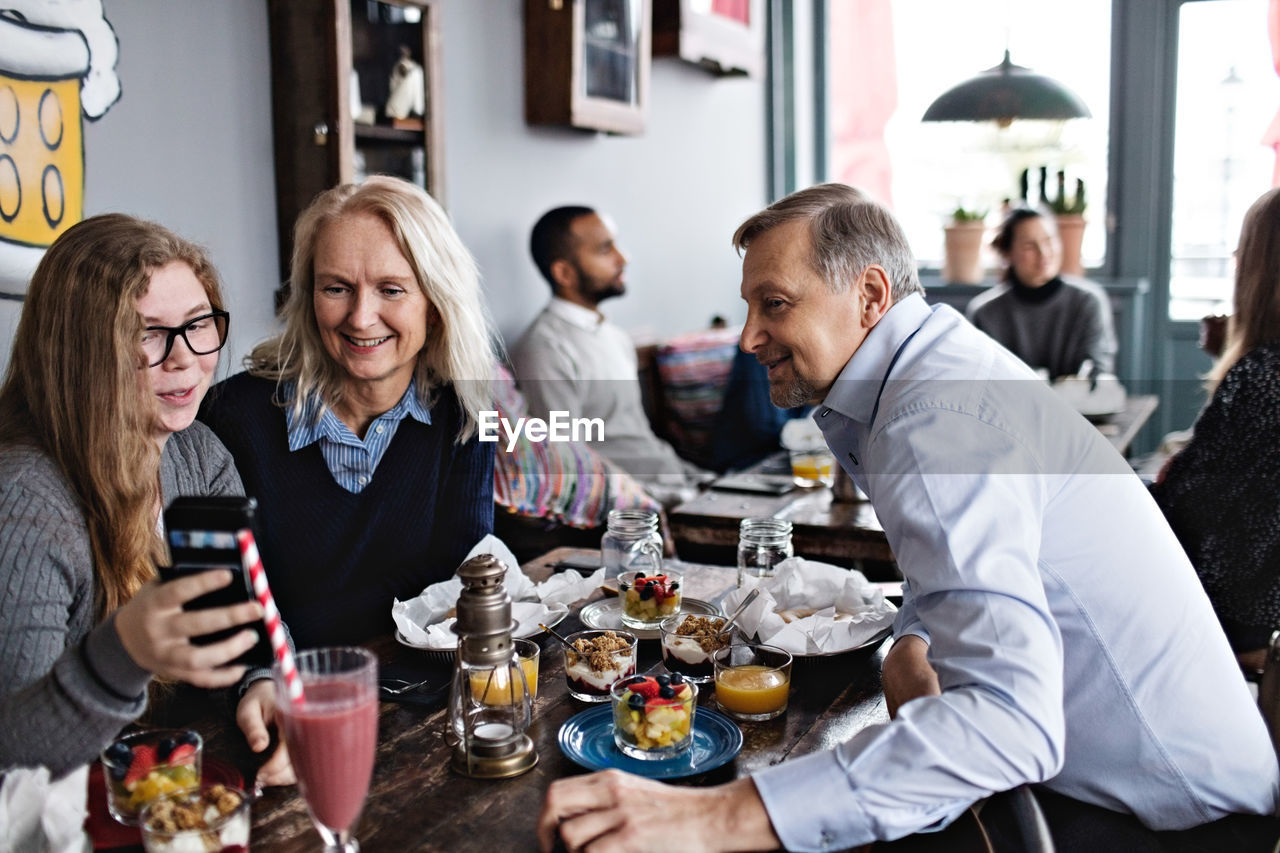 Smiling daughter taking selfie with parents while sitting at table in restaurant