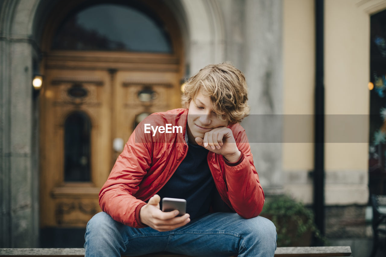 Teenage boy using mobile phone while sitting on bench in city