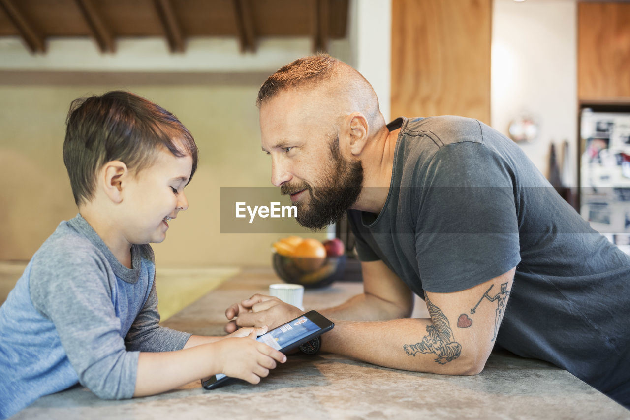 Father looking at smiling son using digital tablet at counter in kitchen