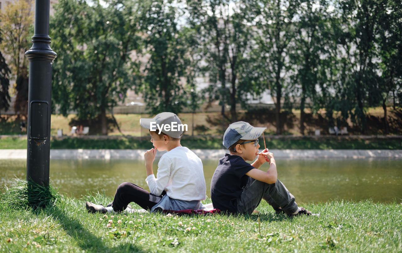 Two boys sit on the grass in the park with their backs to each other and look at the lake. 