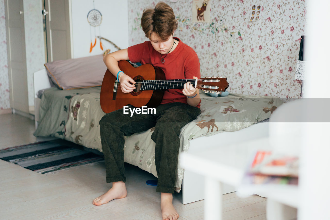 A teenage girl plays the acoustic guitar while sitting in her room.