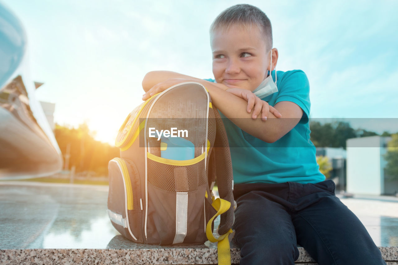 Boy, schoolboy in protective mask sits and wait near school in sunny september day.