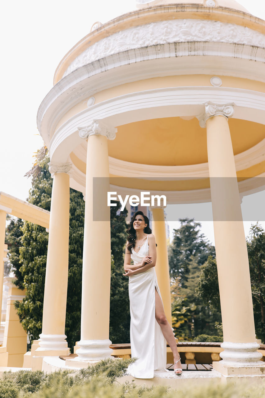 A beautiful brunette lady in an elegant wedding dress poses among the columns in the old city park