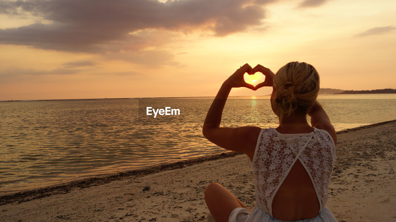 Woman making heart shape at beach during sunset