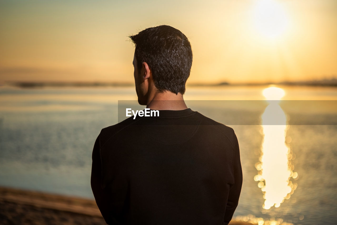 REAR VIEW OF MAN STANDING ON BEACH AGAINST SKY DURING SUNSET