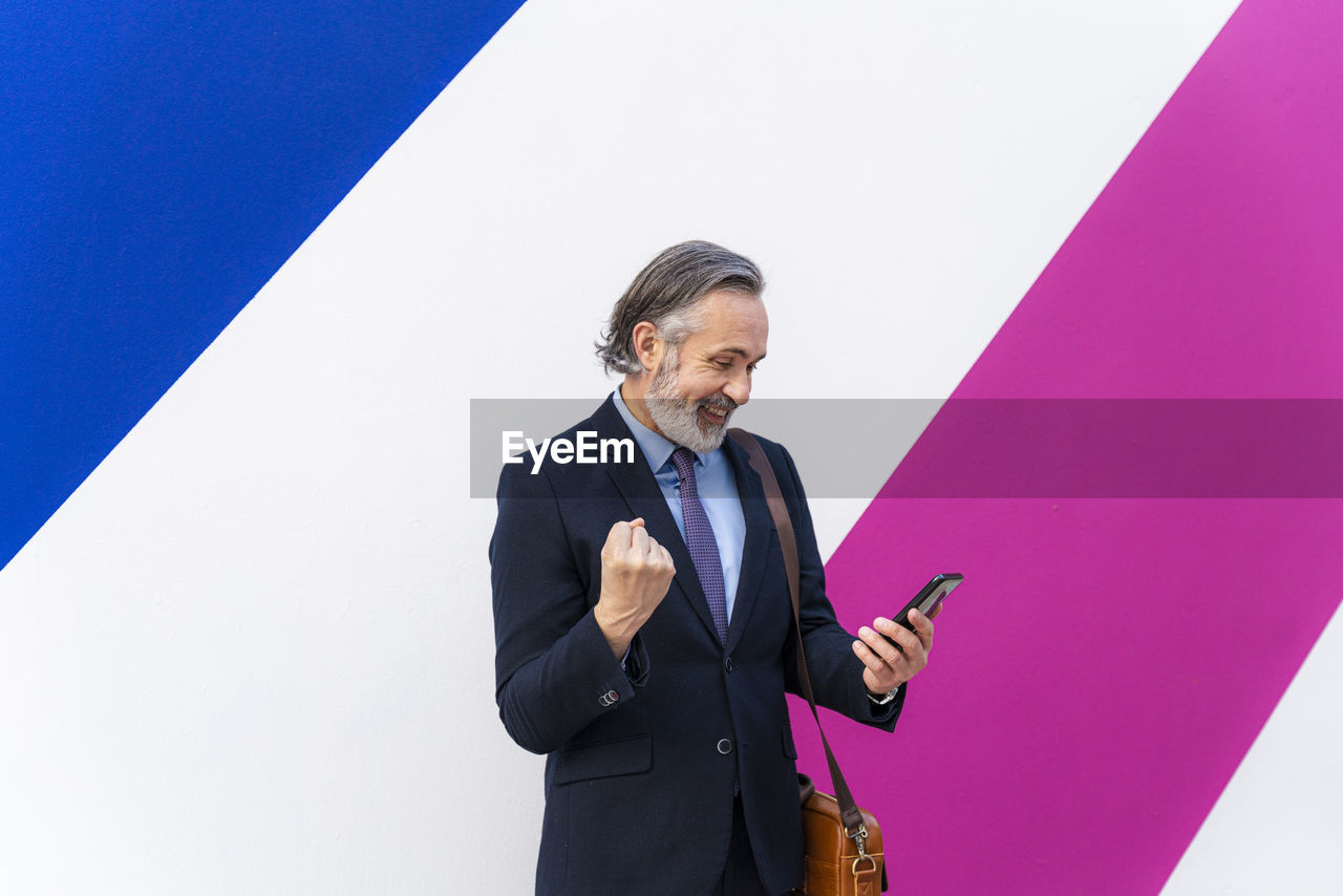 Happy businessman gesturing fist using smart phone standing in front of multi colored wall