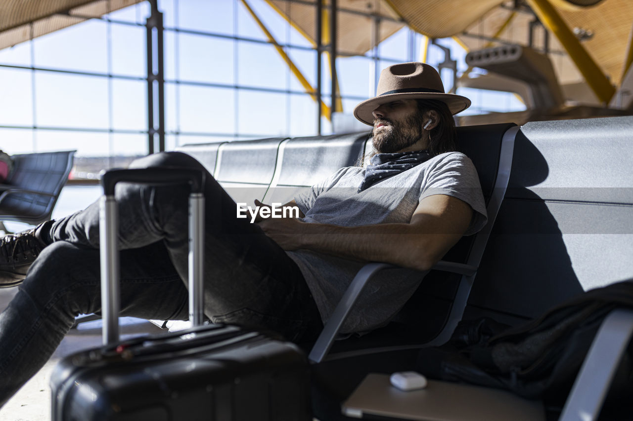 The guy with a hat at the airport in the waiting room sitting waiting for his flight, with wireless headphones to listen to music, sleeps and with his hat covers his eyes