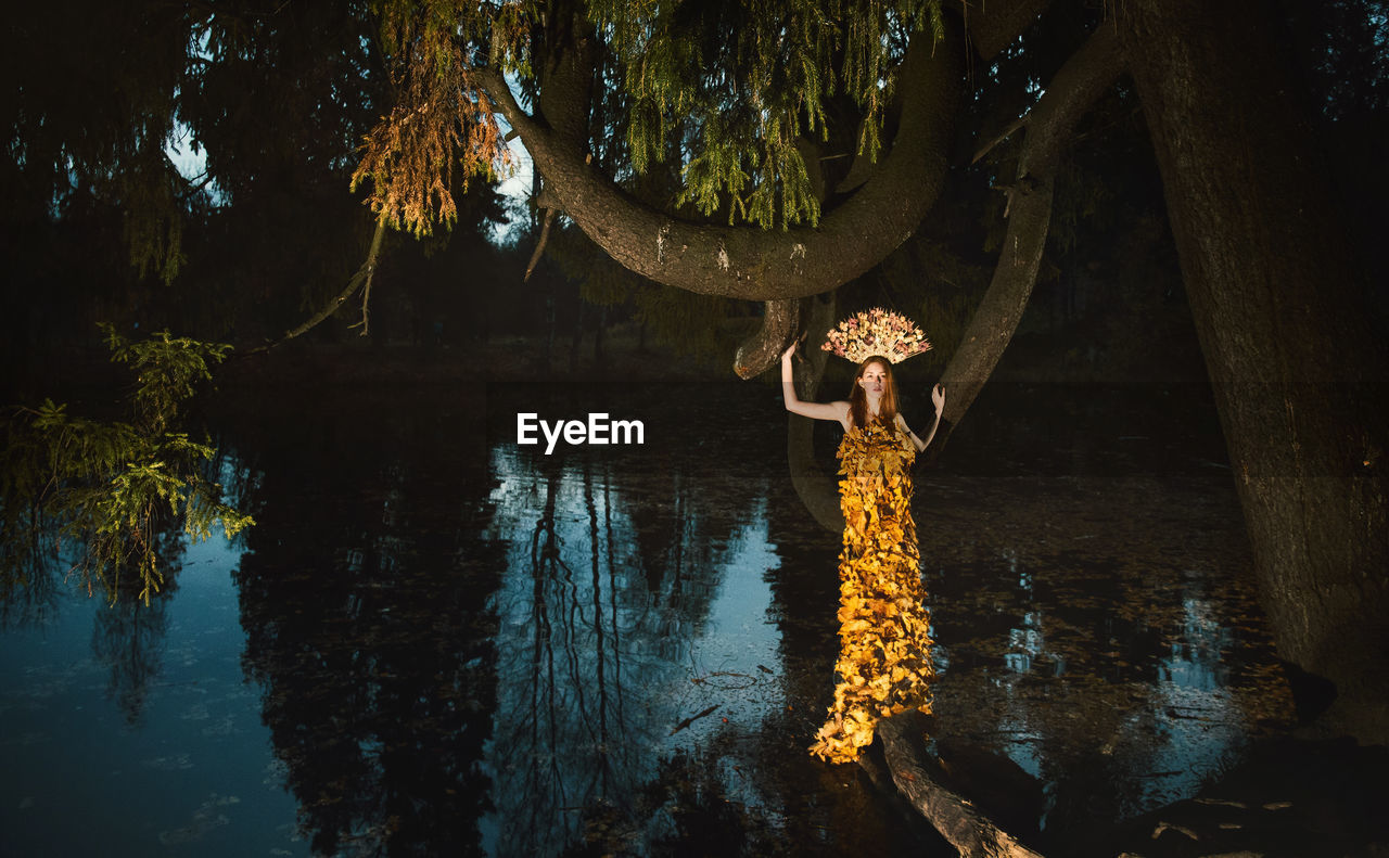 Young woman covered with leaves standing amidst lake at forest during autumn