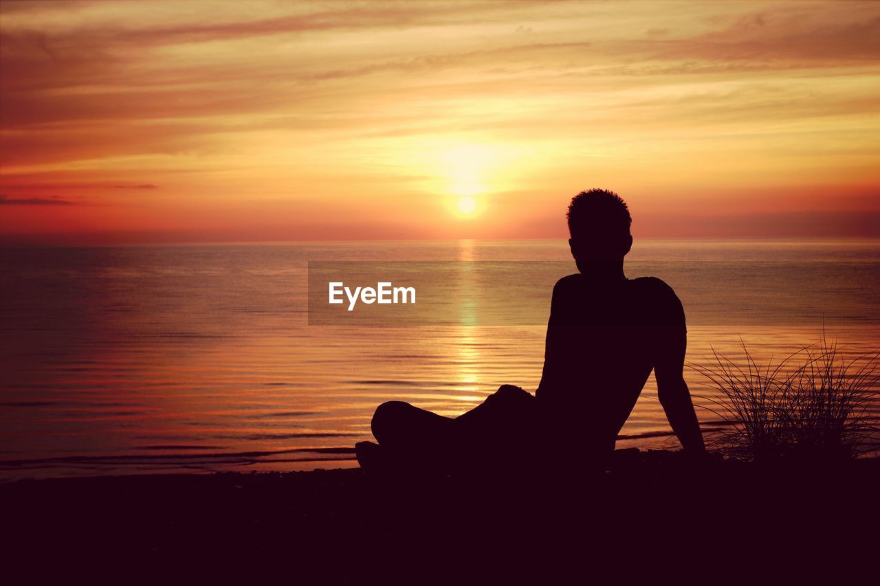 Silhouette man sitting on sea shore during sunset