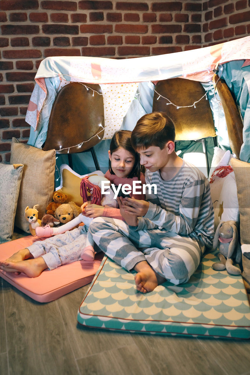 Happy little girl embracing rag doll while looking boy playing video game with phone sitting on tent