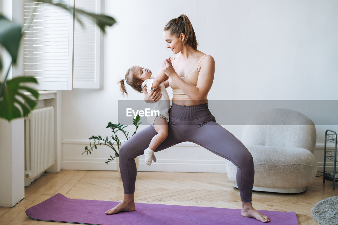 Young woman fit mom with baby girl in her arms doing fitness on mat at home