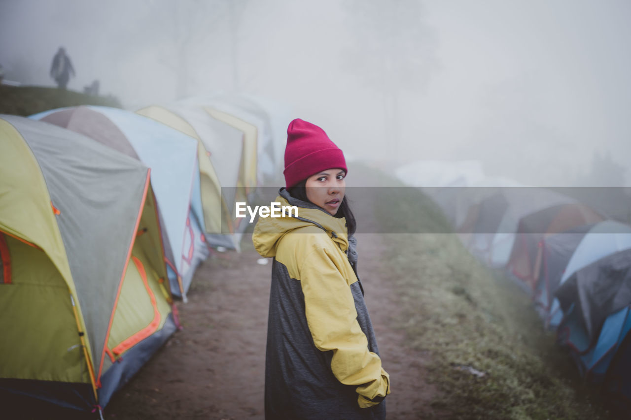 Portrait of woman wearing warm clothing while standing amidst tents during foggy weather