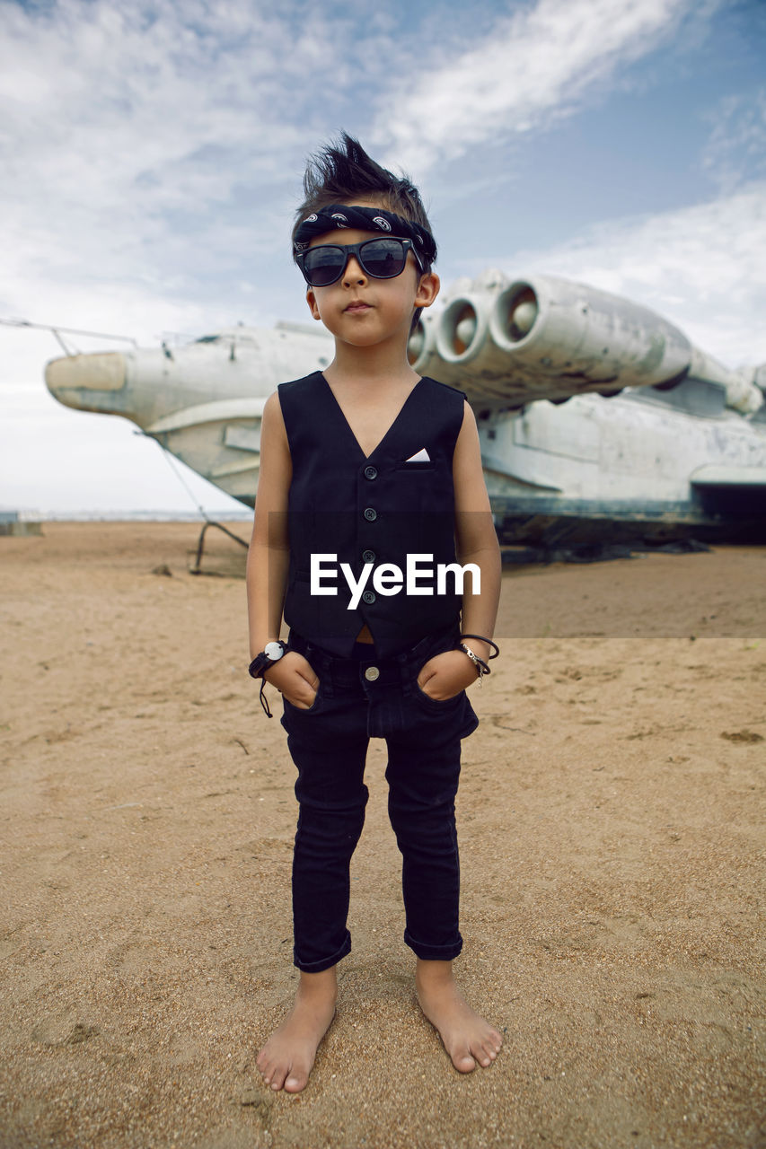 Rocker boy a child in black clothes stands against the abandoned plane on beach in summer