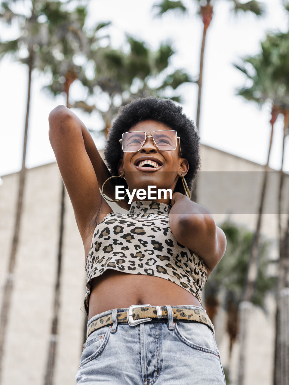 From below of positive young curly haired african american female in casual outfit and trendy sunglasses standing against blurred building on street