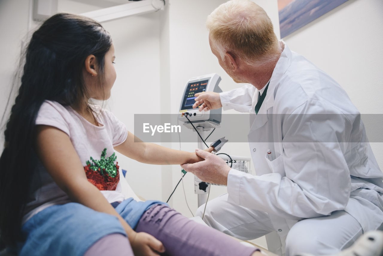 Mature doctor pointing at computer monitor to girl during pulse test in hospital