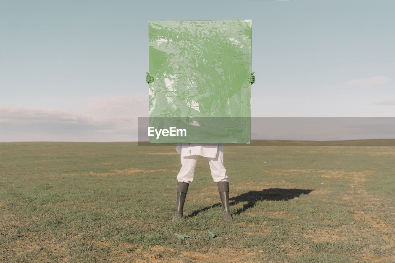 Young woman hiding behind green painting on dry field