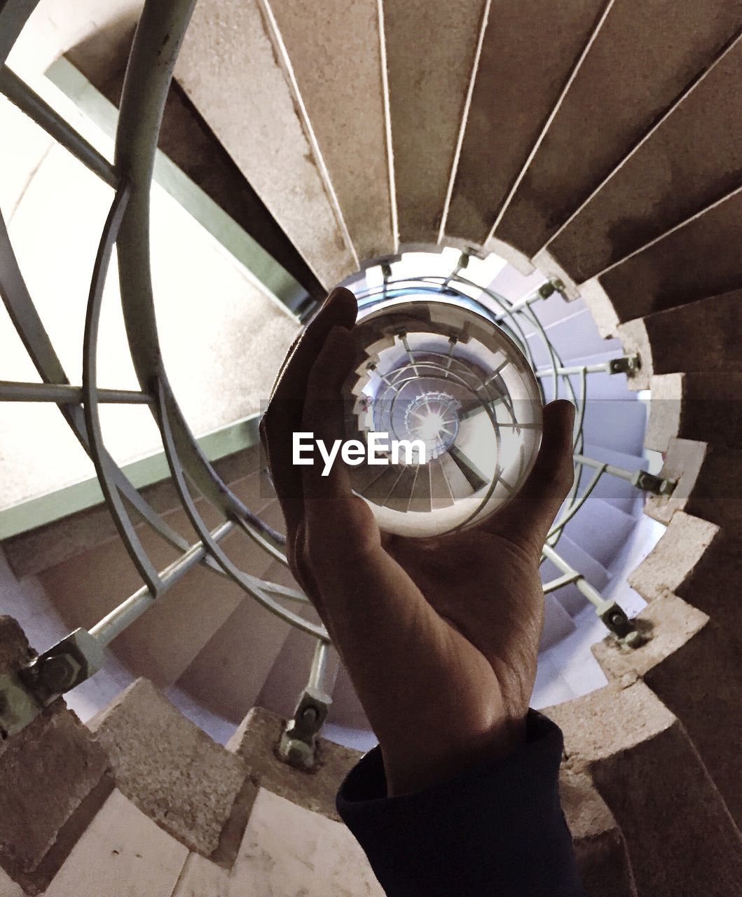 Low angle view of person holding crystal ball with reflection under spiral staircase