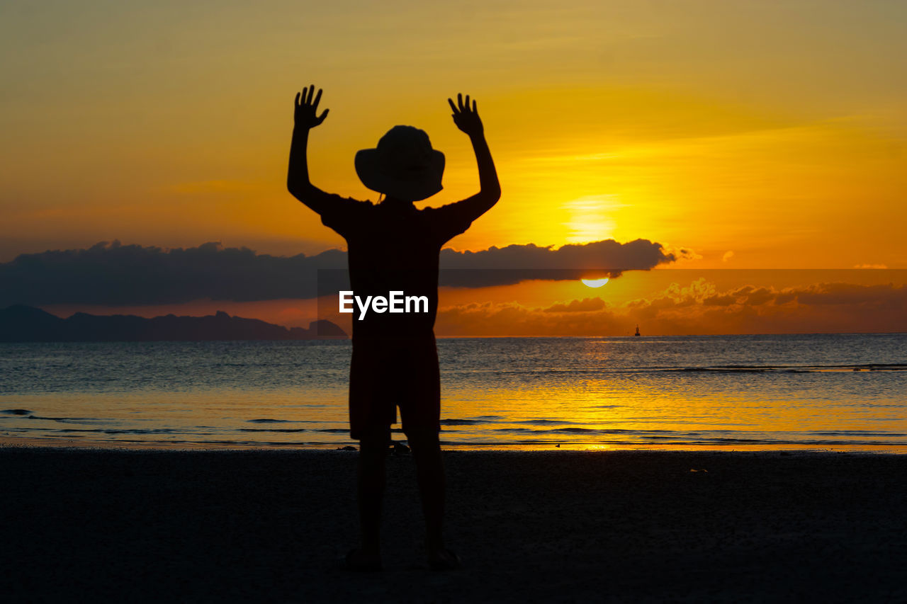 Rear view of silhouette boy with arms raised standing at beach during sunset