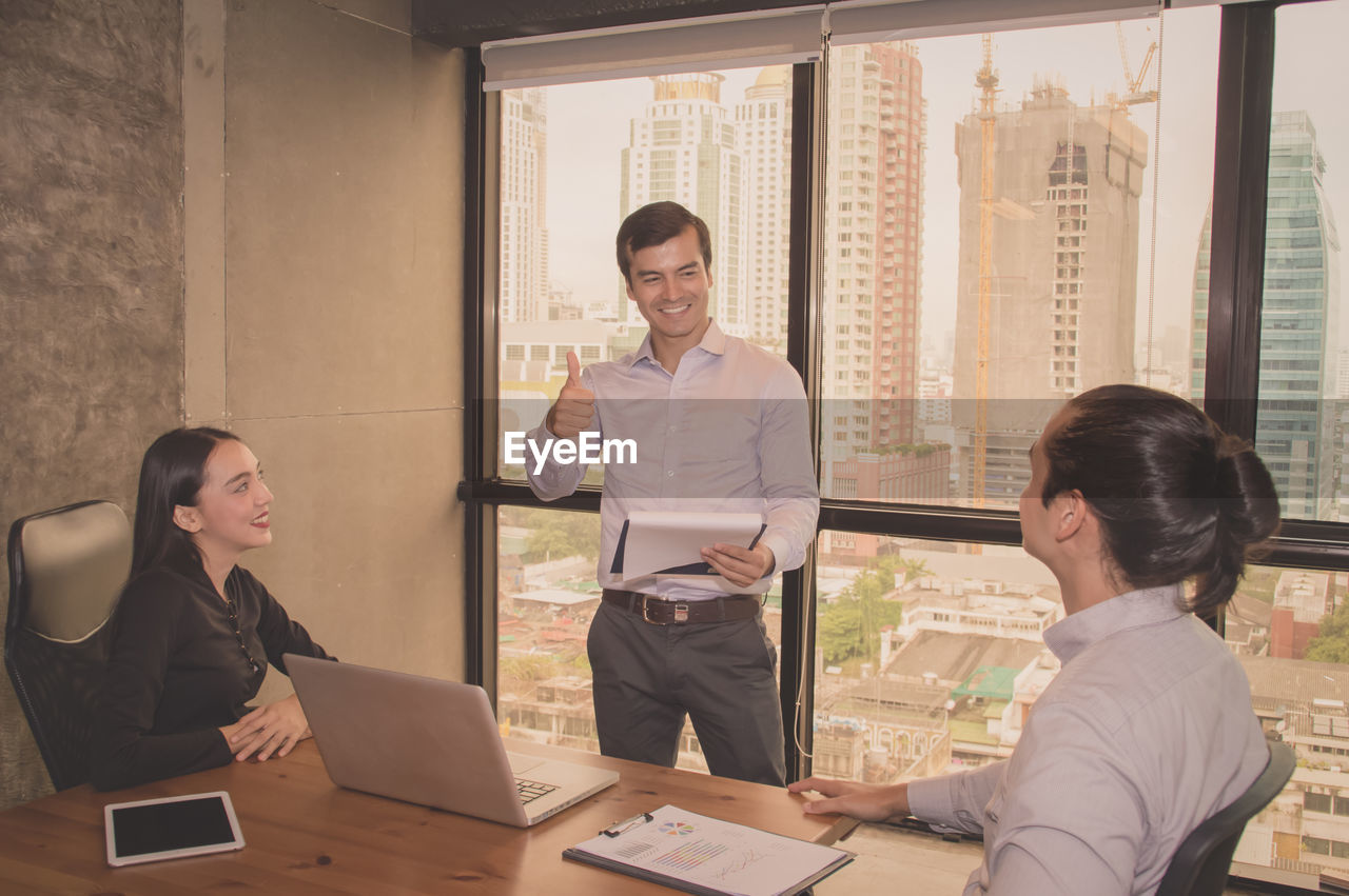 Smiling businessman gesturing thumbs up while discussing with colleagues during meeting in office