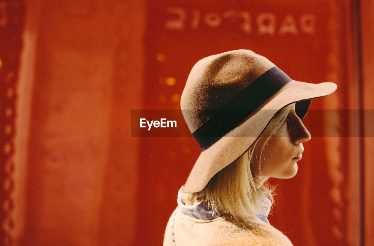 PORTRAIT OF WOMAN WEARING HAT STANDING AGAINST WALL