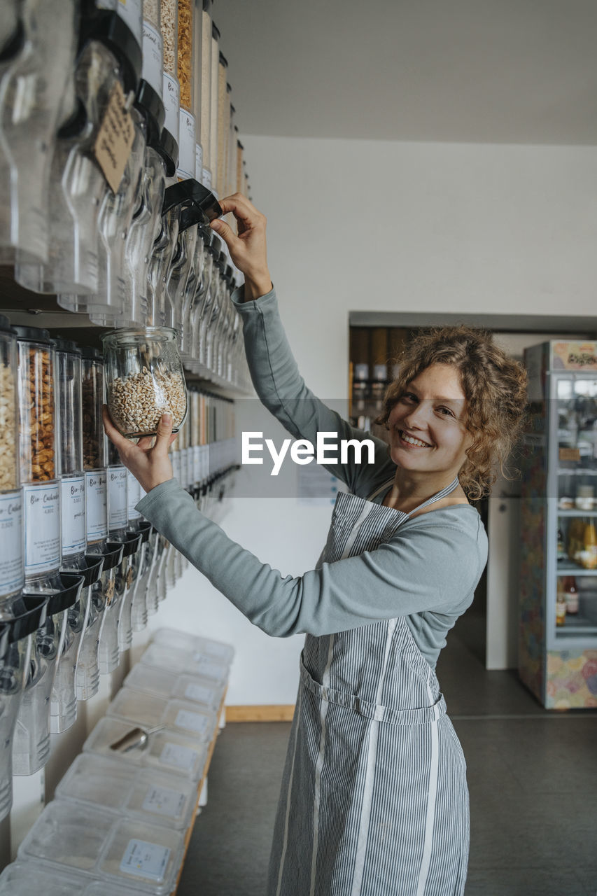 Smiling female store clerk using food dispenser while working in zero waste store