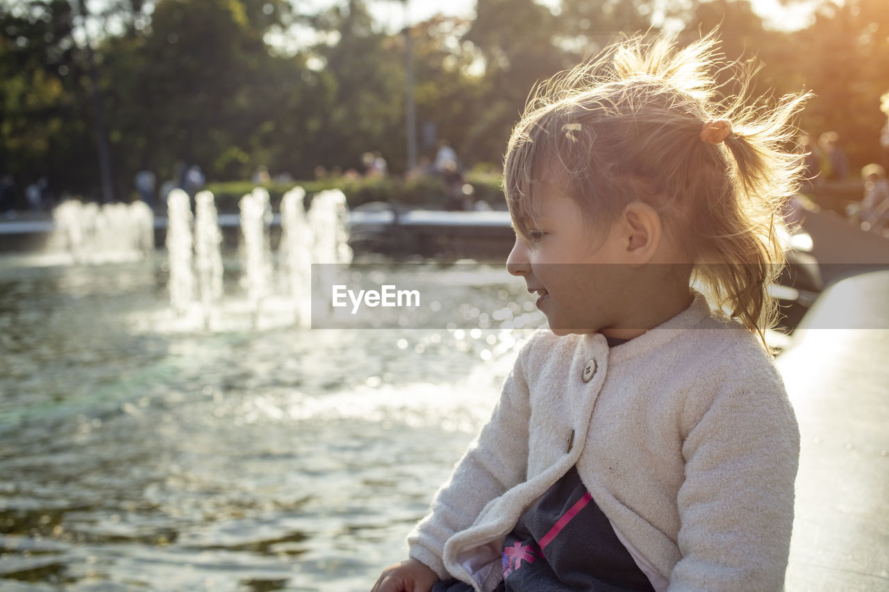 Adorable toddler girl looks at the fountains in the park in the park on a sunny day