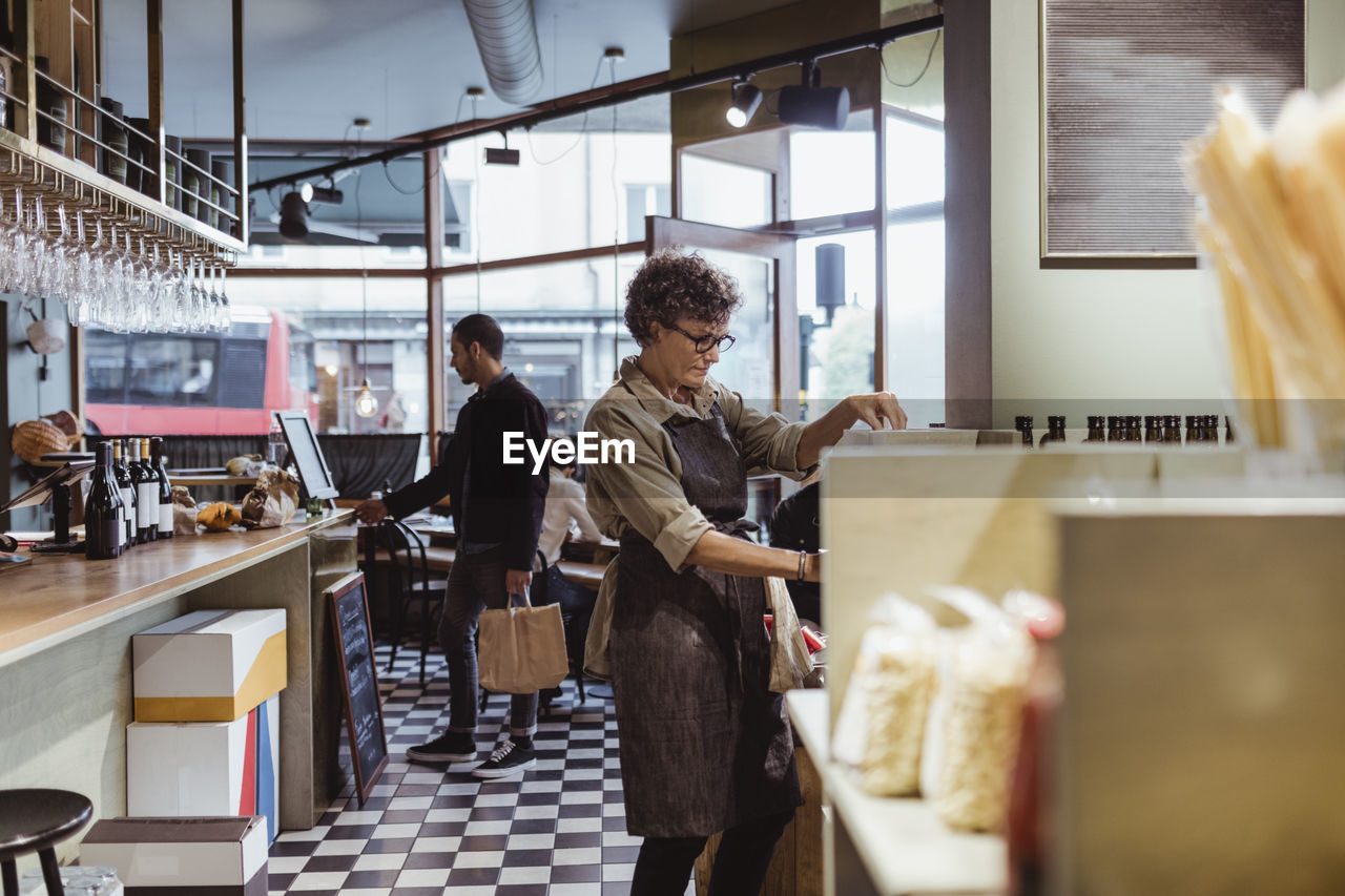 Female store owner working in deli store with customers in background