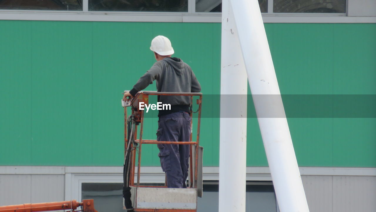 Rear view of construction worker standing on cherry picker against building