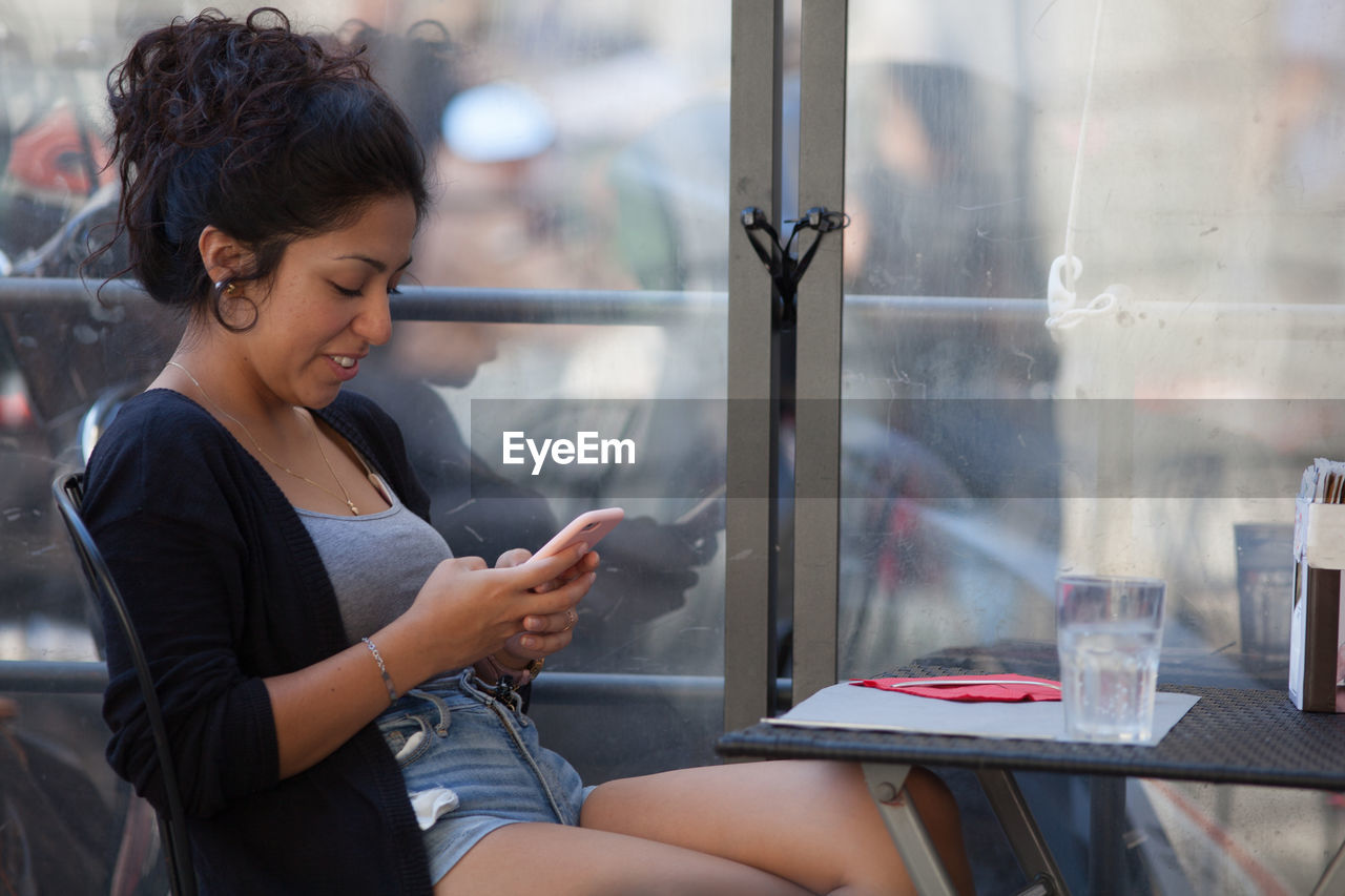 Young woman using mobile phone while sitting by window at restaurant