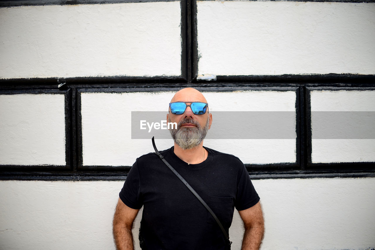 Portrait of a bearded man in black with sunglasses standing against a black and white wall