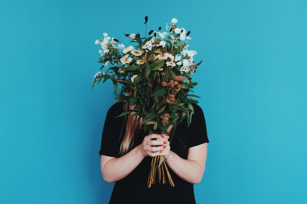 Woman holding bouquet in front of face while standing against blue wall