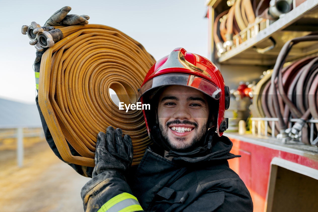 Brave male firefighter wearing protective hardhat and uniform looking at camera while carrying big heavy hose on shoulder near fire truck in farmland