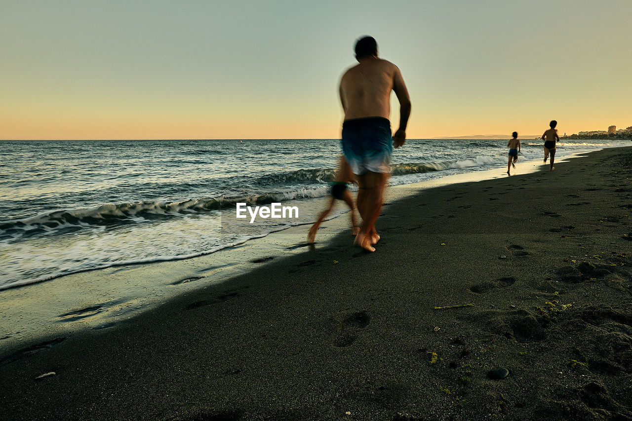FULL LENGTH OF SHIRTLESS MAN ON BEACH AGAINST SKY
