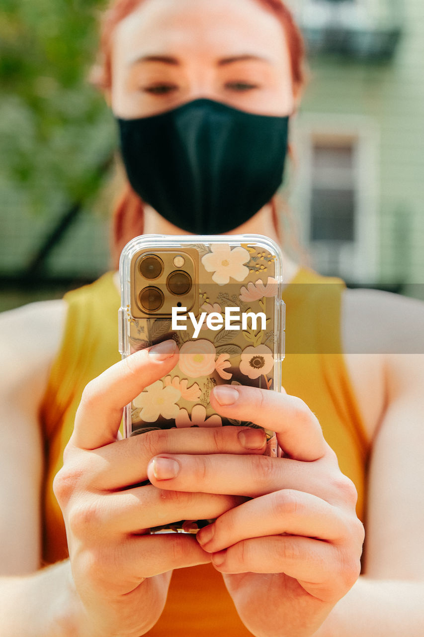 Young woman sitting outdoors texting on phone, wearing face mask.