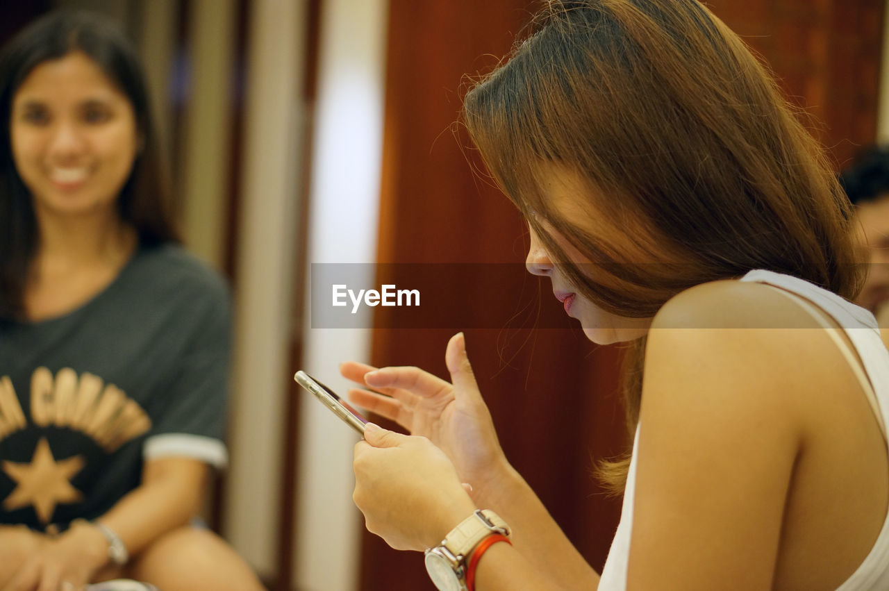 Side view of young woman using phone with friend in background