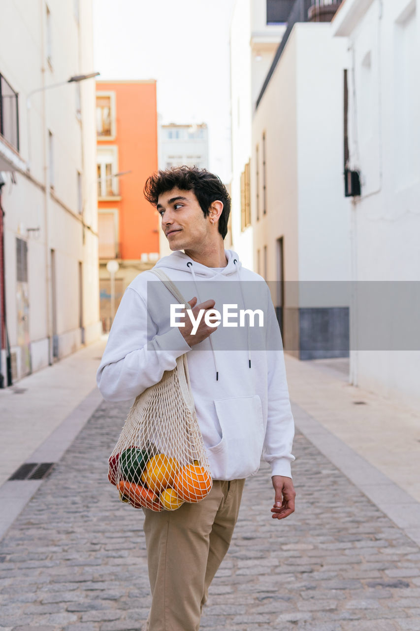 Side view of content male standing with eco friendly mesh bag full of fresh fruits in city street looking away