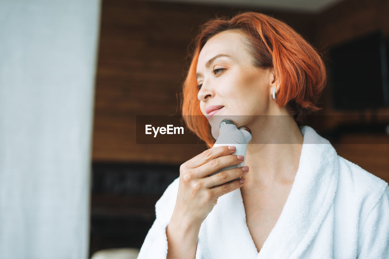 Woman in white bathrobe with red hair doing fasial massage with microcurrent facial massager