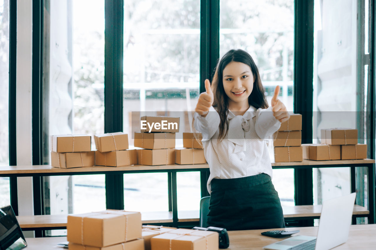 Portrait of smiling young woman showing thumbs up signs by cardboard boxes at office