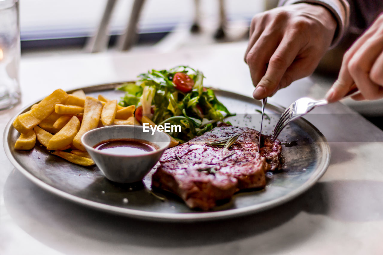 Close-up of hands cutting into steak served on plate with fries