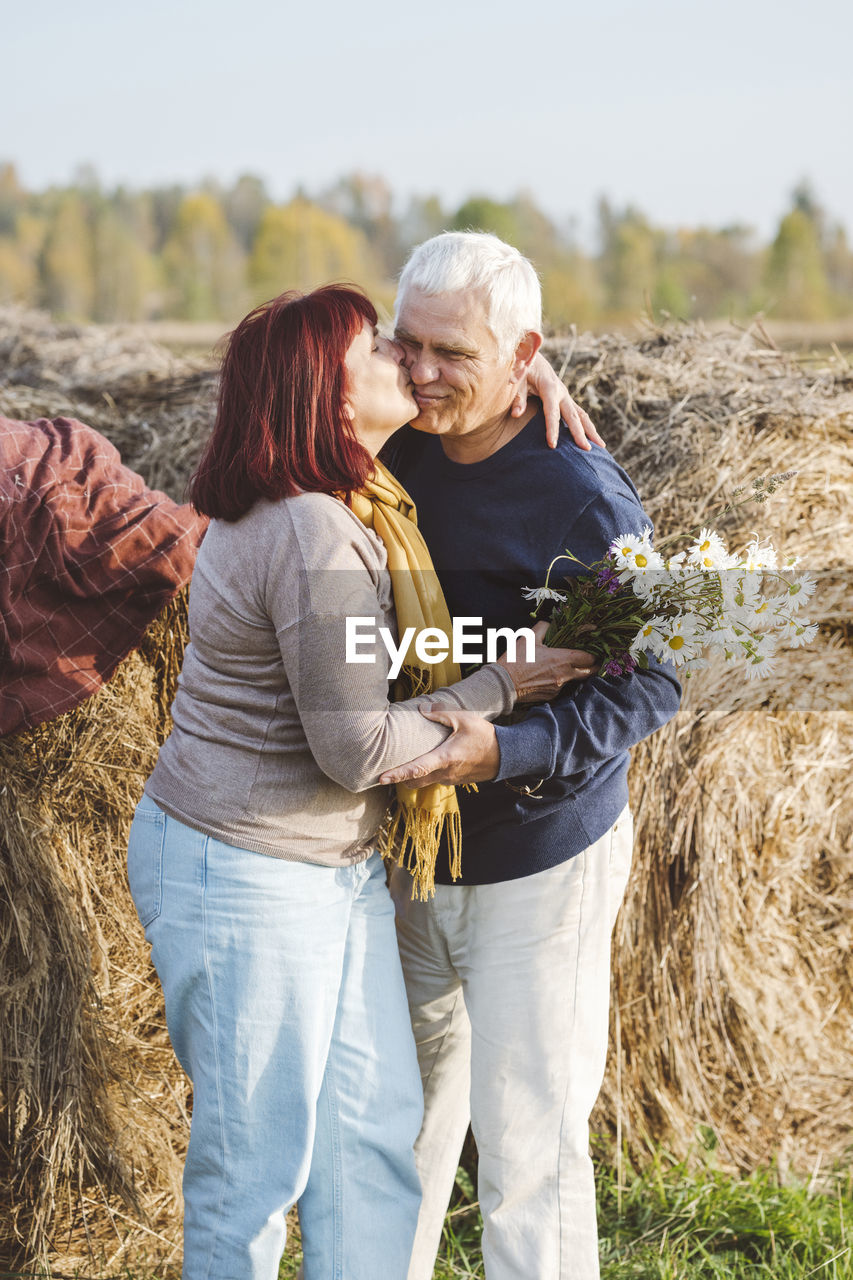 Happy senior couple kissing while standing against hay bales at field