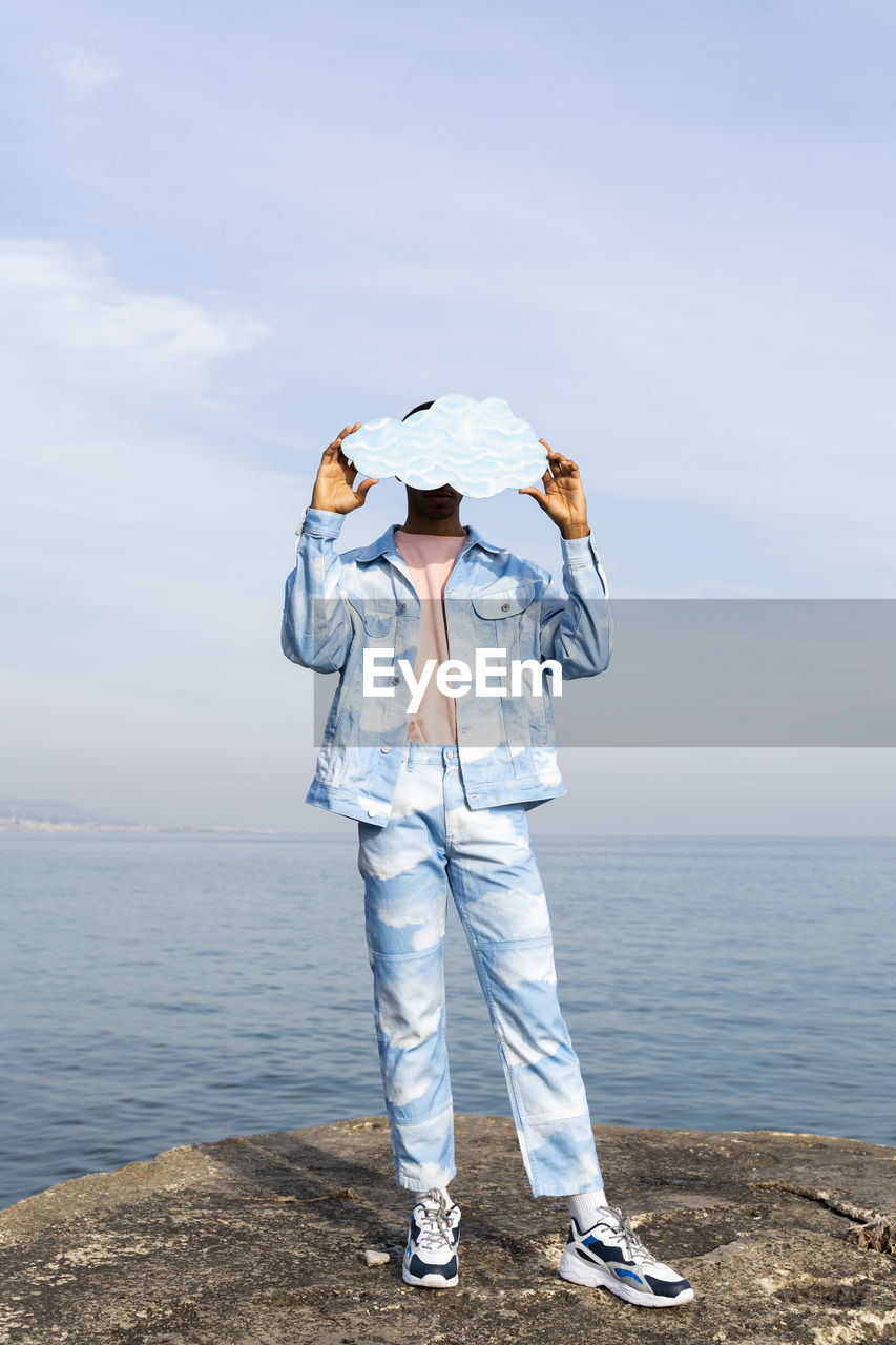 Young man hiding face with cloud cut out while standing against sea