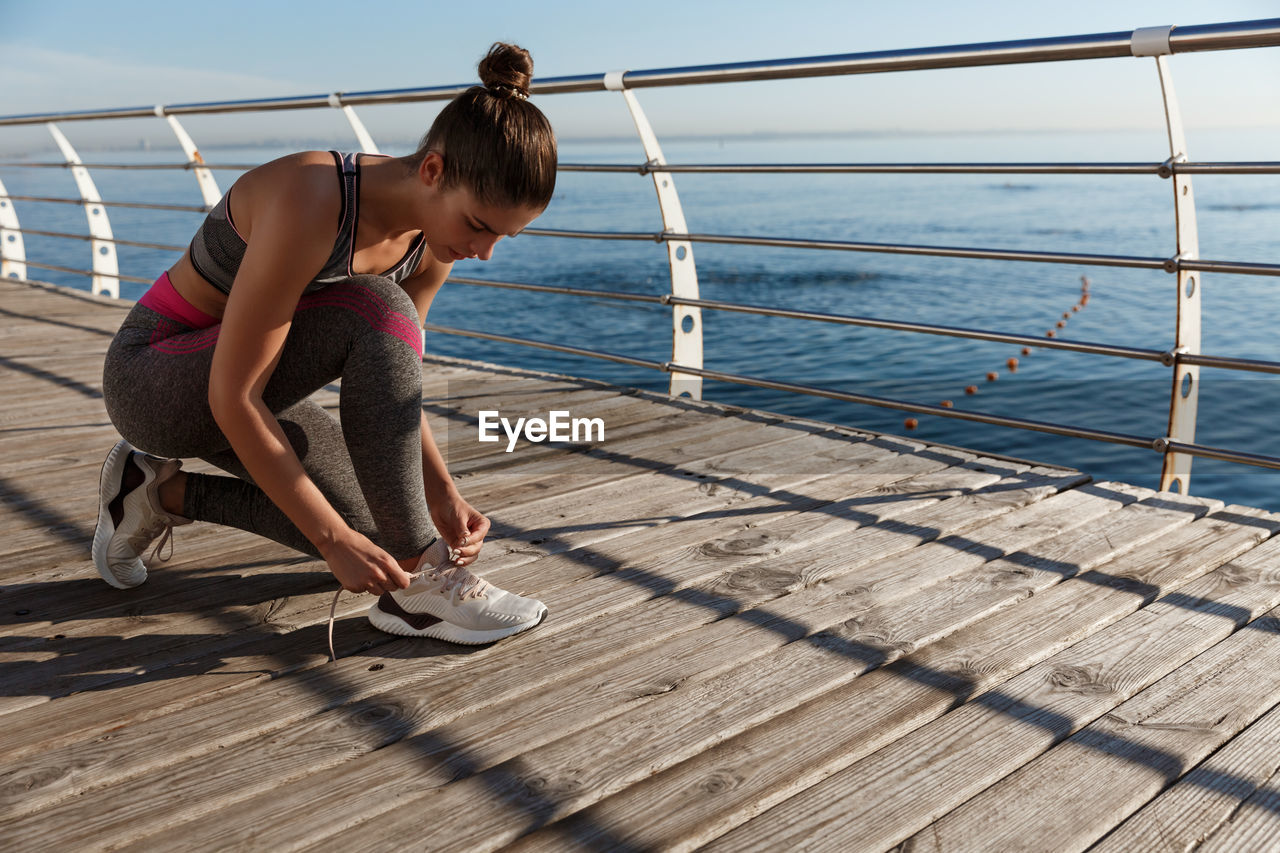 Full length of woman tying shoelace on floor against sea and sky