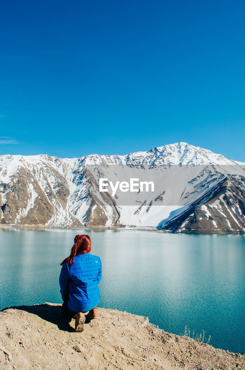 Rear view of young woman crouching by lake against mountain range