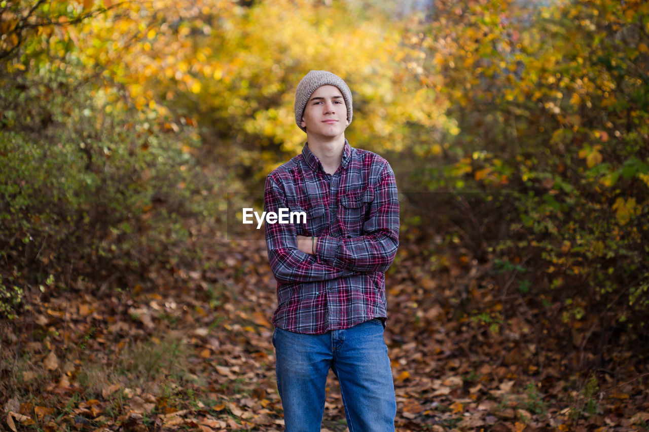 Confident teen boy stands in woods alone with arms crossed.